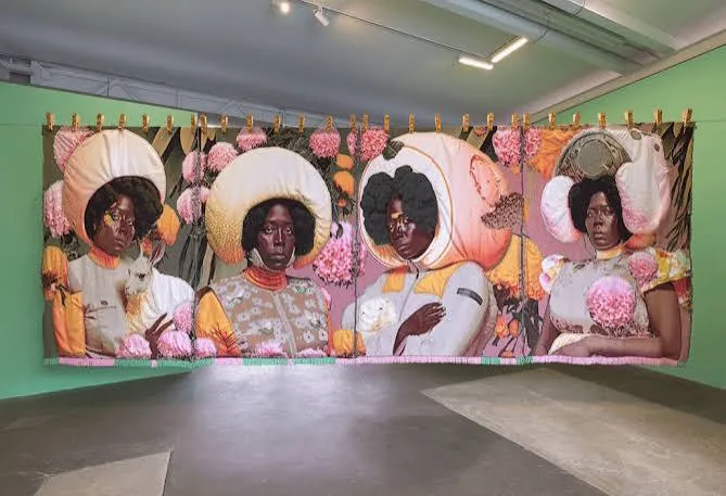 A large textile artwork of four women in bright headdresses, hanging in a gallery